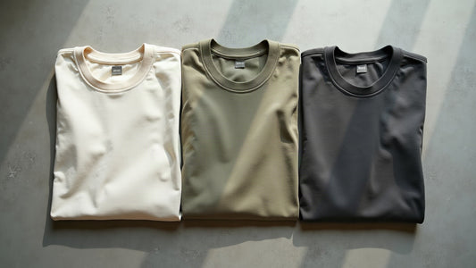 Flat lay of folded garment-dyed crew neck cotton t-shirts in ecru, sage and charcoal in soft daylight.