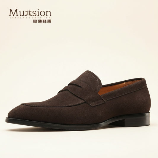 Espresso brown suede penny loafer with tonal saddle strap and welted sole on a warm off-white studio backdrop.