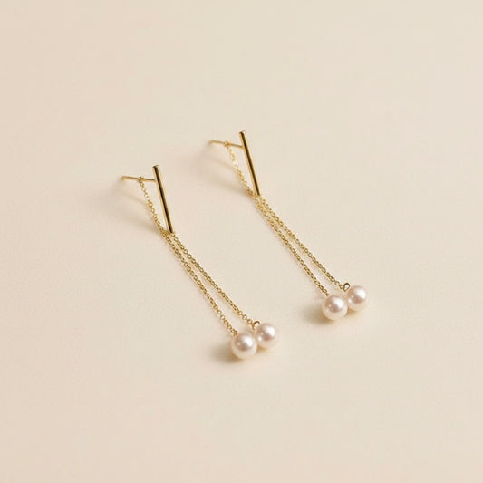 Fine gold threader drop earrings with twin Akoya pearls, styled on warm off-white textured surface in soft natural light.