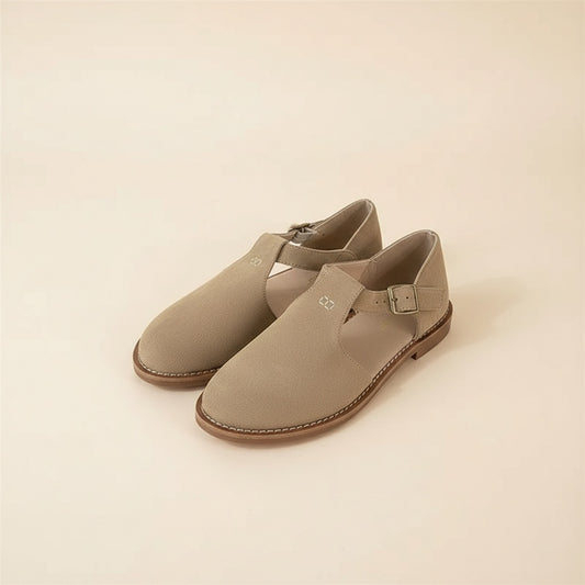 Taupe suede closed-toe T-bar flat sandals with side buckle, shown as a pair on a warm textured neutral surface.