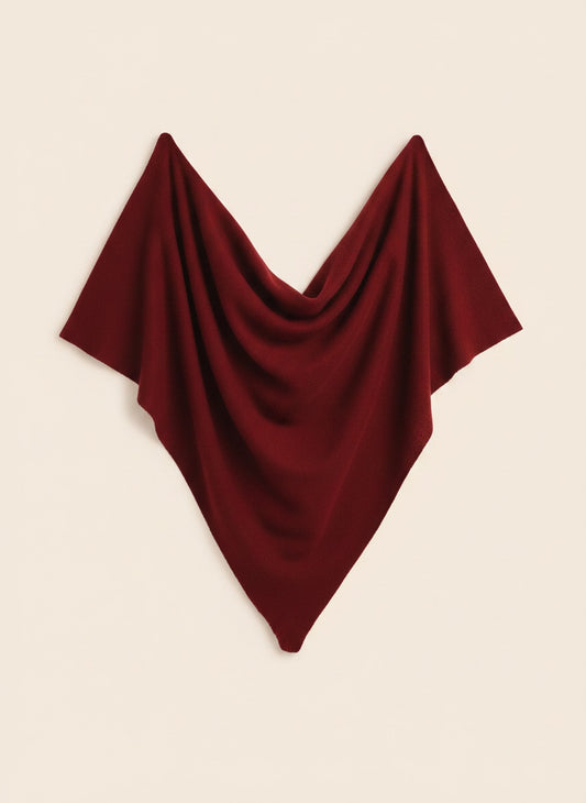Deep burgundy triangular wool scarf softly draped against a warm off-white wall, tapered point falling naturally beside a ceramic vase.