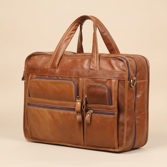 Tan full-grain leather briefcase with twin rolled handles, front zip pockets and brass D-ring, styled on warm neutral linen.
