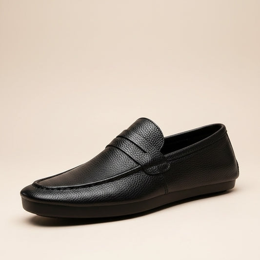 Black pebbled leather penny loafer with tonal strap and low rubber sole on warm travertine ledge.