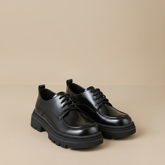 Chunky black leather platform derby shoes with lugged track sole, styled on a warm off-white plinth in soft directional light.