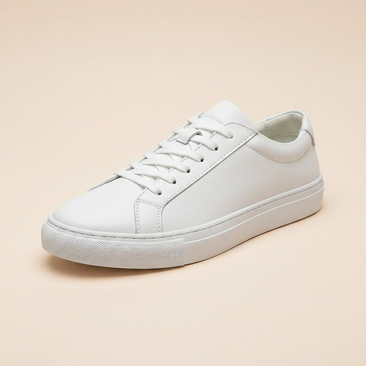 Minimalist white leather low-top court sneaker with tonal rubber sole and flat cotton laces on a warm stone ledge.