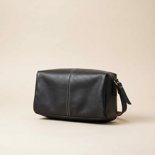 Compact black top-grain leather crossbody bag with tonal centre stitch and slim strap, styled on warm travertine stone.