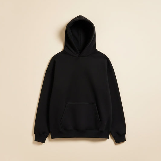 Relaxed black brushed cotton pullover hoodie with kangaroo pocket and soft hood, styled with tonal trousers on a warm studio backdrop.