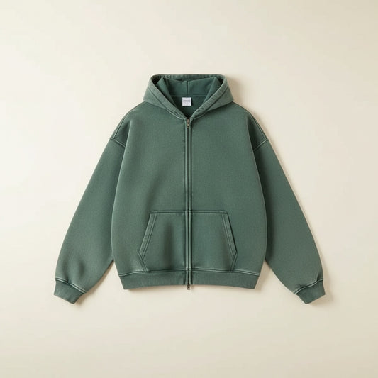 Men's washed sage green full-zip hoodie styled with charcoal trousers and black loafers in a soft, minimal studio setting.