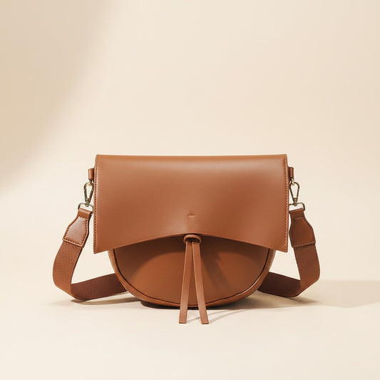 Tan leather foldover crossbody bag with tonal tassel pull and webbing strap on warm off-white stone surface.