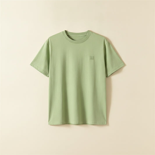 Soft sage green cotton crew neck t-shirt with subtle tonal chest detail, styled with stone tapered trousers in warm studio light.