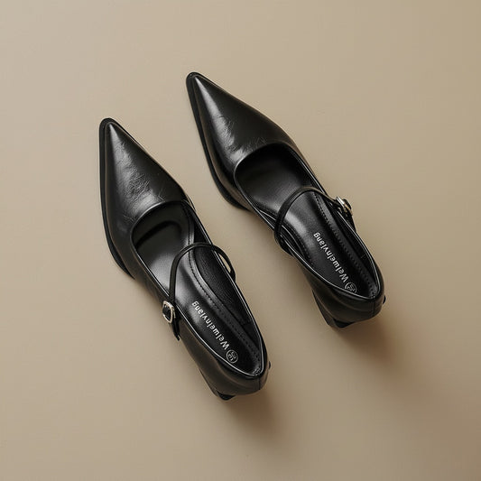Pointed-toe black leather Mary Jane flats with slim strap and polished buckle on warm off-white studio backdrop.