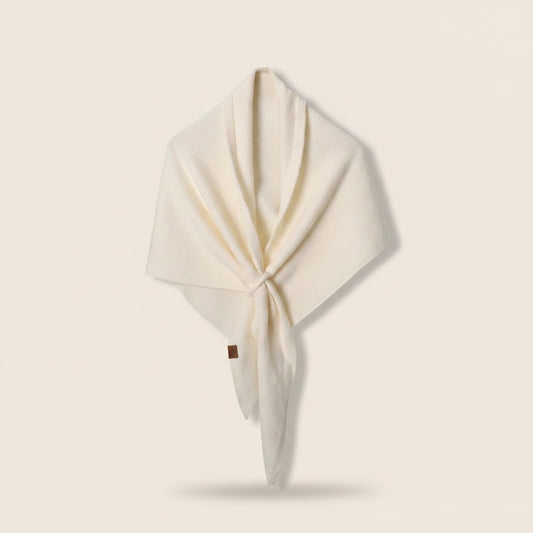 Ivory wool-blend triangle shawl with soft front knot and leather tab, styled on a warm travertine ledge against a plaster wall.