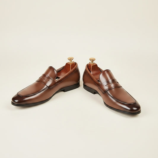 Chestnut brown leather penny loafers with almond toe and low stacked heel, styled on warm off-white studio backdrop.