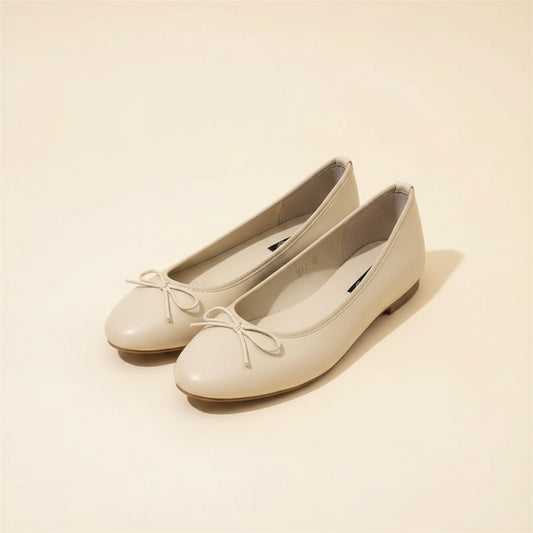 Cream leather ballet flats with slim tonal bow and soft rounded toe, styled on a warm off-white studio backdrop.