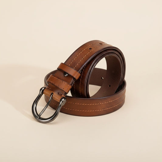 Coiled tan full-grain leather pin belt with curved gunmetal buckle and tonal centre stitching on a warm off-white studio backdrop.