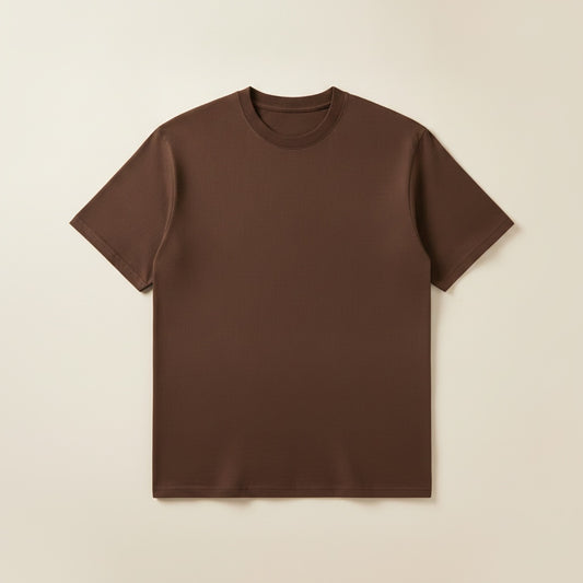 Man in deep chocolate brown heavyweight cotton crewneck t-shirt, styled with charcoal trousers against a warm neutral wall.