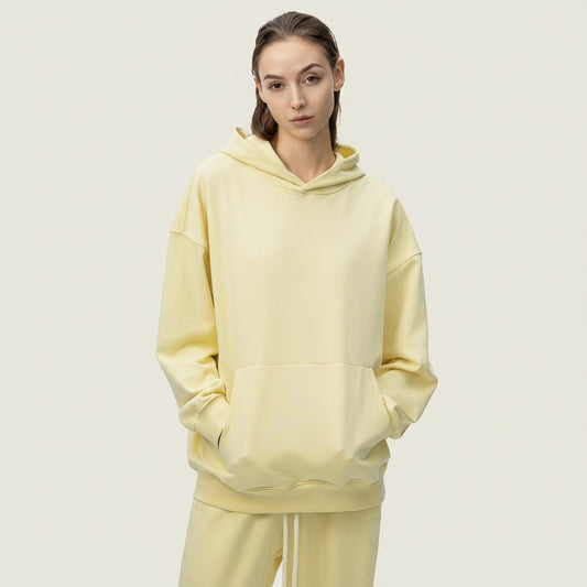 Woman in pale butter-yellow oversized loopback hoodie with kangaroo pocket, styled with tonal cream trousers on warm off-white studio backdrop.