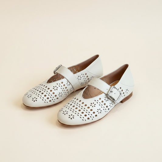 Ivory perforated leather Mary Jane flats with floral laser-cut detailing and silver buckle strap on warm neutral backdrop.