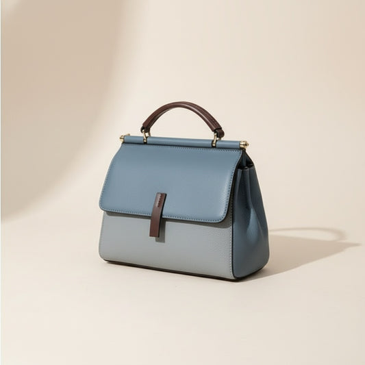 Structured dusty blue leather top-handle satchel with burgundy tab closure, styled on a travertine plinth in warm daylight.