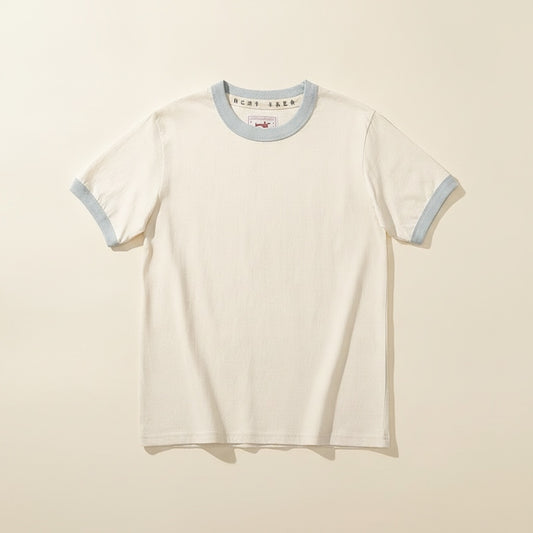 Ivory ringer crew t-shirt with pale blue rib trim, worn tucked into tailored trousers in a quiet editorial studio setting.