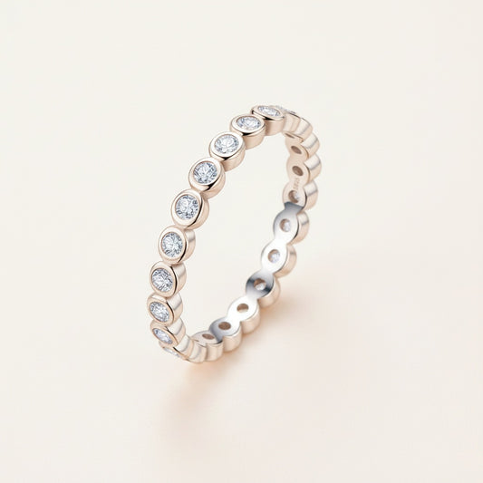Slim sterling silver bezel-set moissanite eternity ring standing on a warm stone plinth in soft natural light.