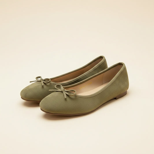Sage suede ballet flats with delicate tonal bow and almond toe, styled on warm off-white surface in soft daylight.
