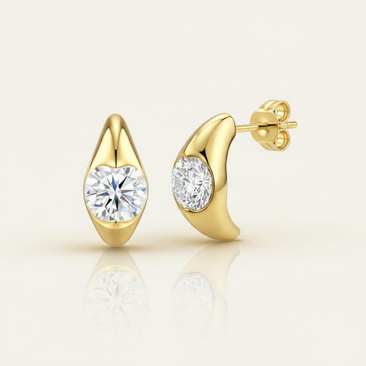 Marquise-shaped gold-tone stud earrings with bezel-set round moissanite stones on a warm neutral surface in soft daylight.