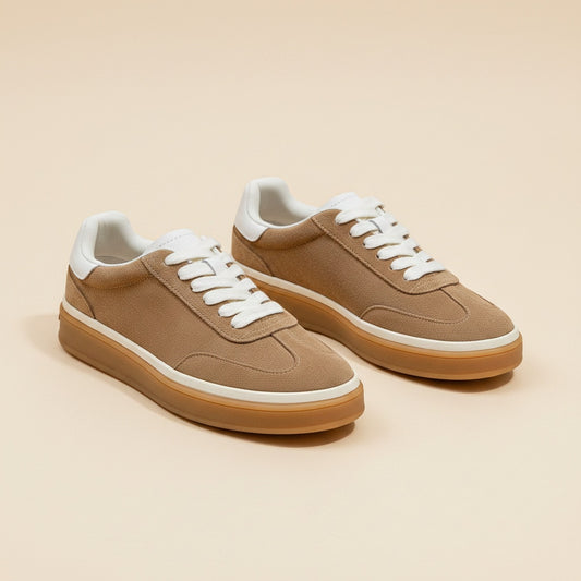 Camel low-top court trainers with cream laces, white heel tab and gum sole on a warm travertine stone surface.