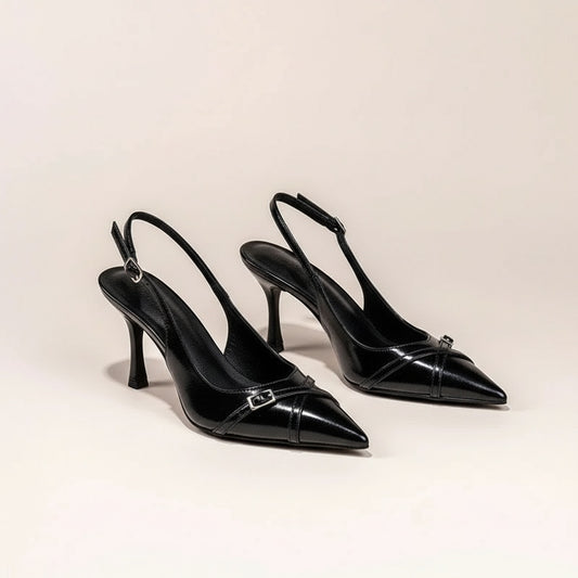Black pointed-toe slingback pumps with sculpted stiletto heel and slim buckle strap, styled on warm neutral stone.