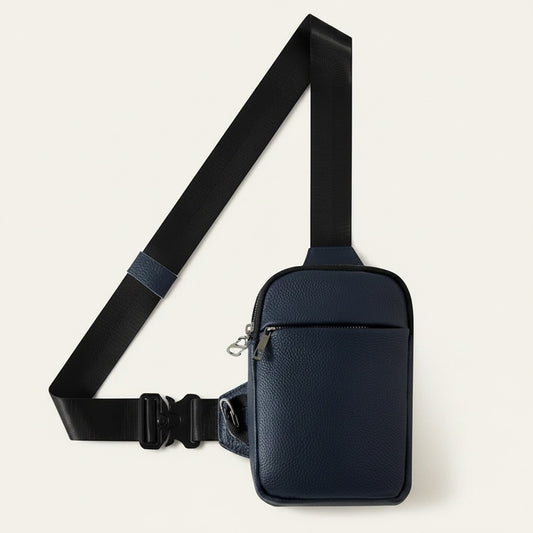 Navy pebbled-leather crossbody sling bag with black webbing strap and front zip pocket, styled against a warm sunlit wall.