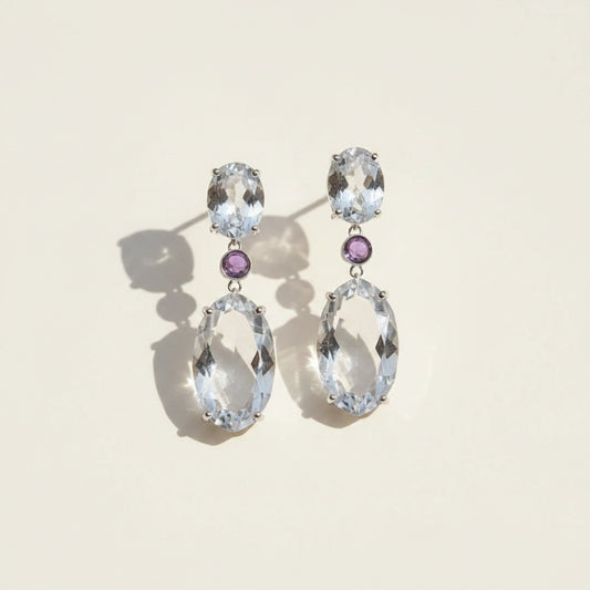Sterling silver drop earrings with faceted oval clear quartz stones and violet amethyst accents on a warm off-white background.
