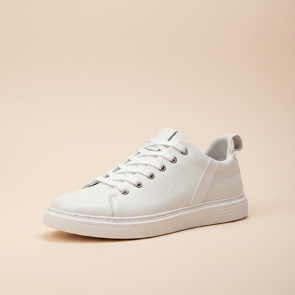 White pebbled leather low-top court sneaker with tonal laces and cupsole, styled on a warm stone plinth in soft daylight.