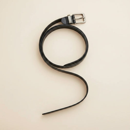Slim black leather belt with minimal brushed-metal square buckle, coiled on a warm off-white studio backdrop.