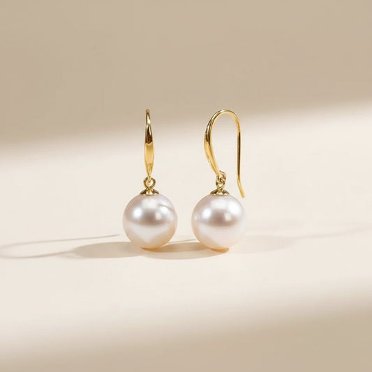Akoya pearl drop earrings with slim 18K yellow gold hooks on a warm neutral stone surface, soft daylight.
