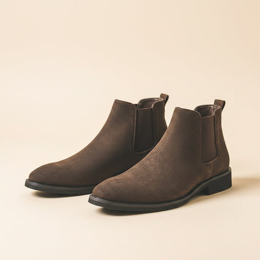 Dark brown suede Chelsea boots with twin elastic gores and low stacked heel on a warm plinth in soft directional light.